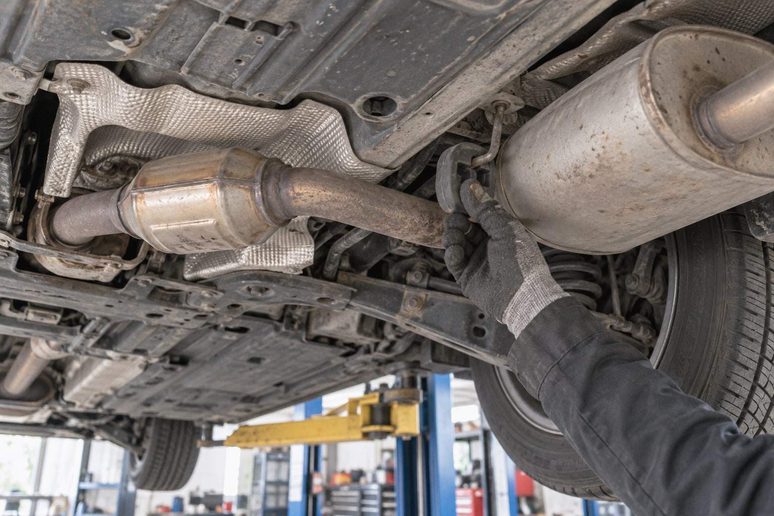 Eine Hand im Arbeitshandschuh prüft die Aufhängung einer Abgasanlage am Unterboden eines Fahrzeugs auf der Hebebühne